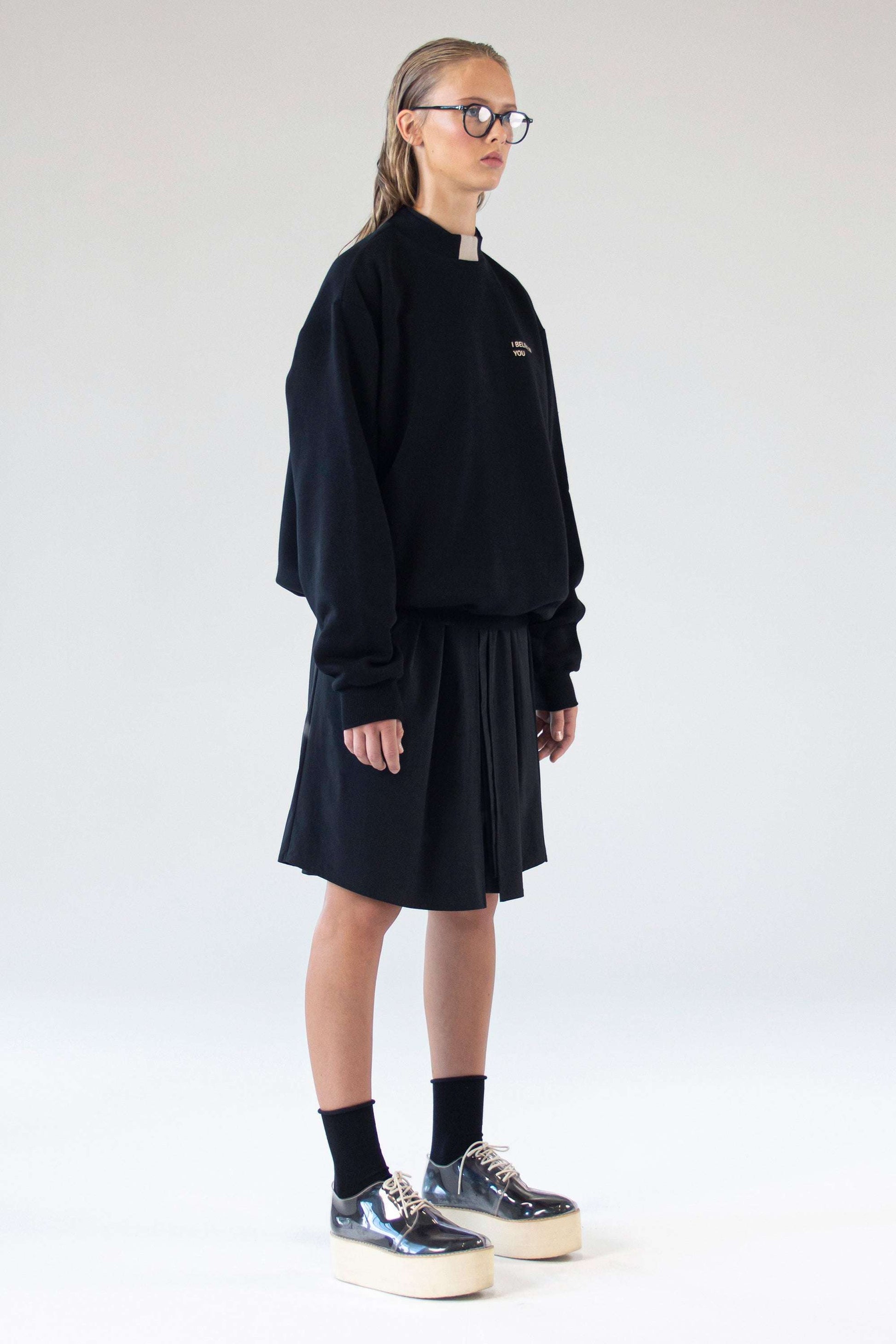 Full-body front view of model wearing One Wolf ‘I Believe In You’ unisex black sweater paired with black skirt.
